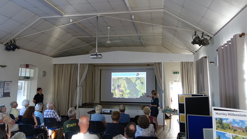 A village hall with a woman at the front pointing to a screen with a map on and a few rows of seated people (by Julie Grainger)