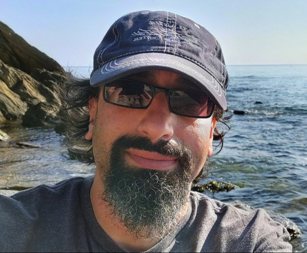 A man with a beard, sunglasses and a hat smiling at the camera with a backdrop of sea and cliffs