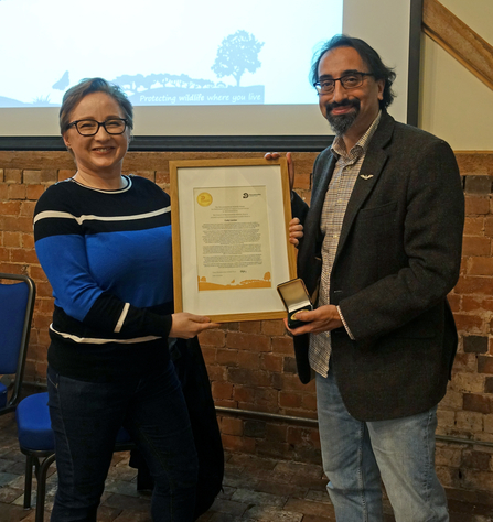 A woman on the left presenting a framed certificate to a man on the right, both are smiling at the camera