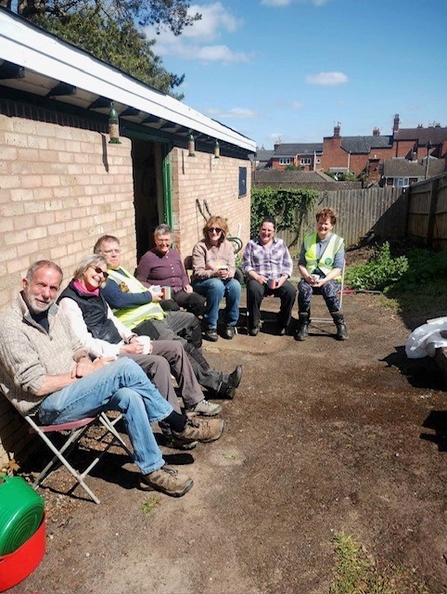 A group of seven people sitting on chairs in a yard on a sunny day - they're smiling and look like they're enjoying the cuppas that they're drinking