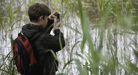 Boy with a backpack on standing by water taking a photograph by Paul Harris/2020VISION